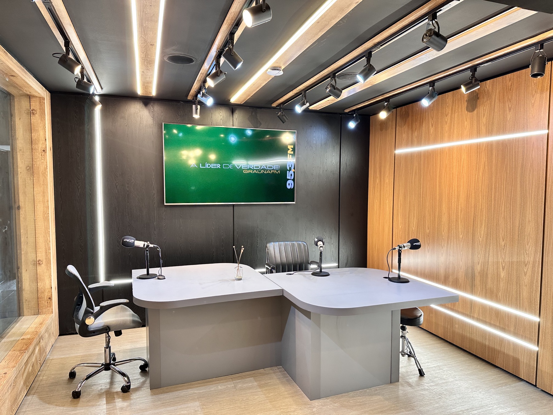 Bancada de locução iluminada com três microfones e painel de vídeo com a marca da Graúna FM
