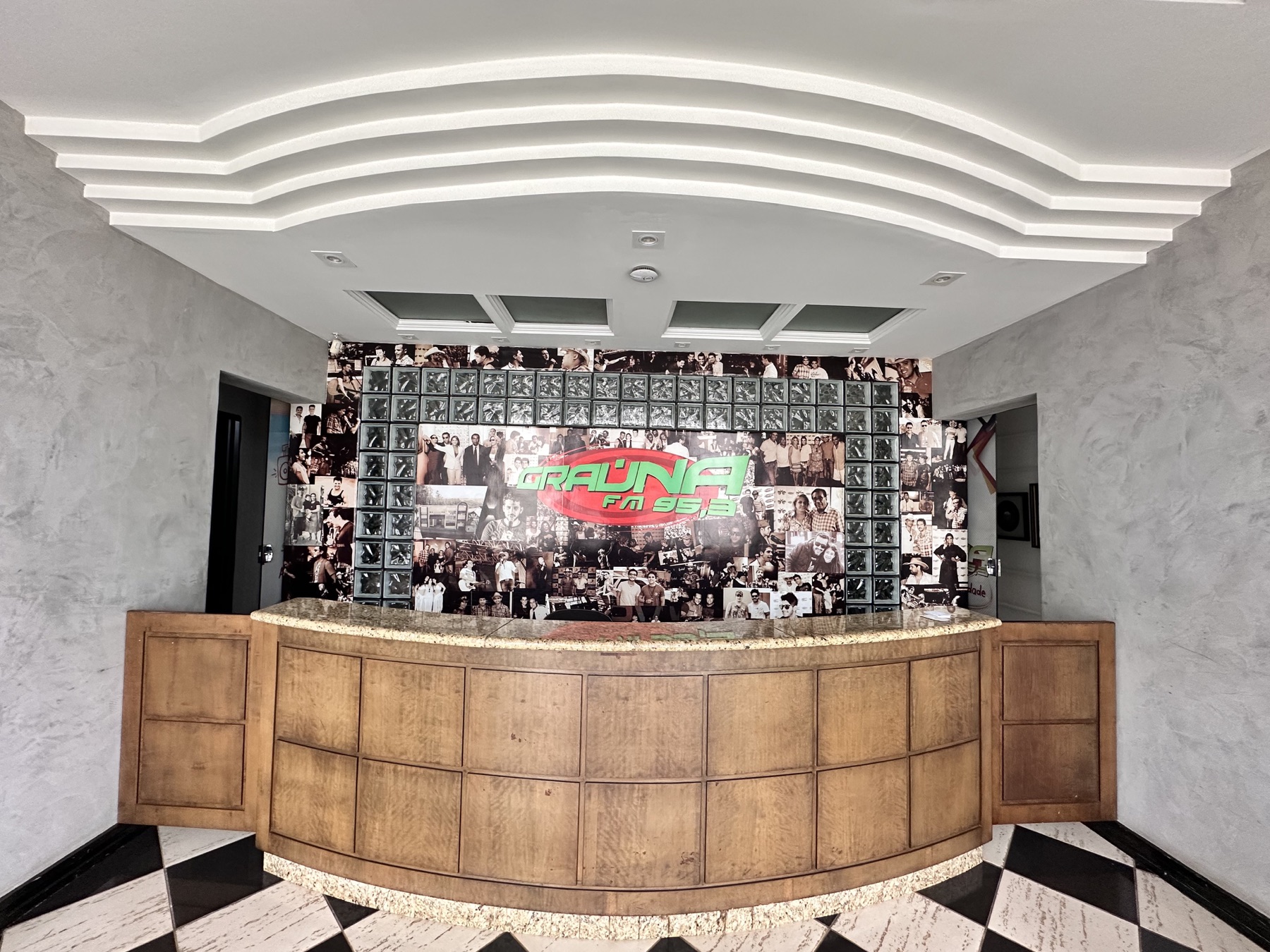 Recepção da Rádio Graúna FM com balcão de madeira curvo e painel de fotos com o logotipo da emissora ao fundo