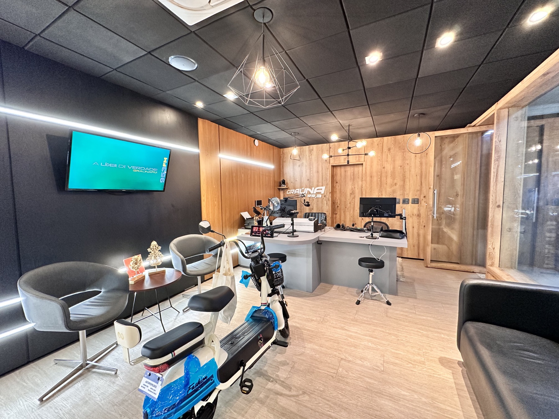 Sala de convivência da rádio com poltronas, televisor da emissora e bancada de operação ao fundo