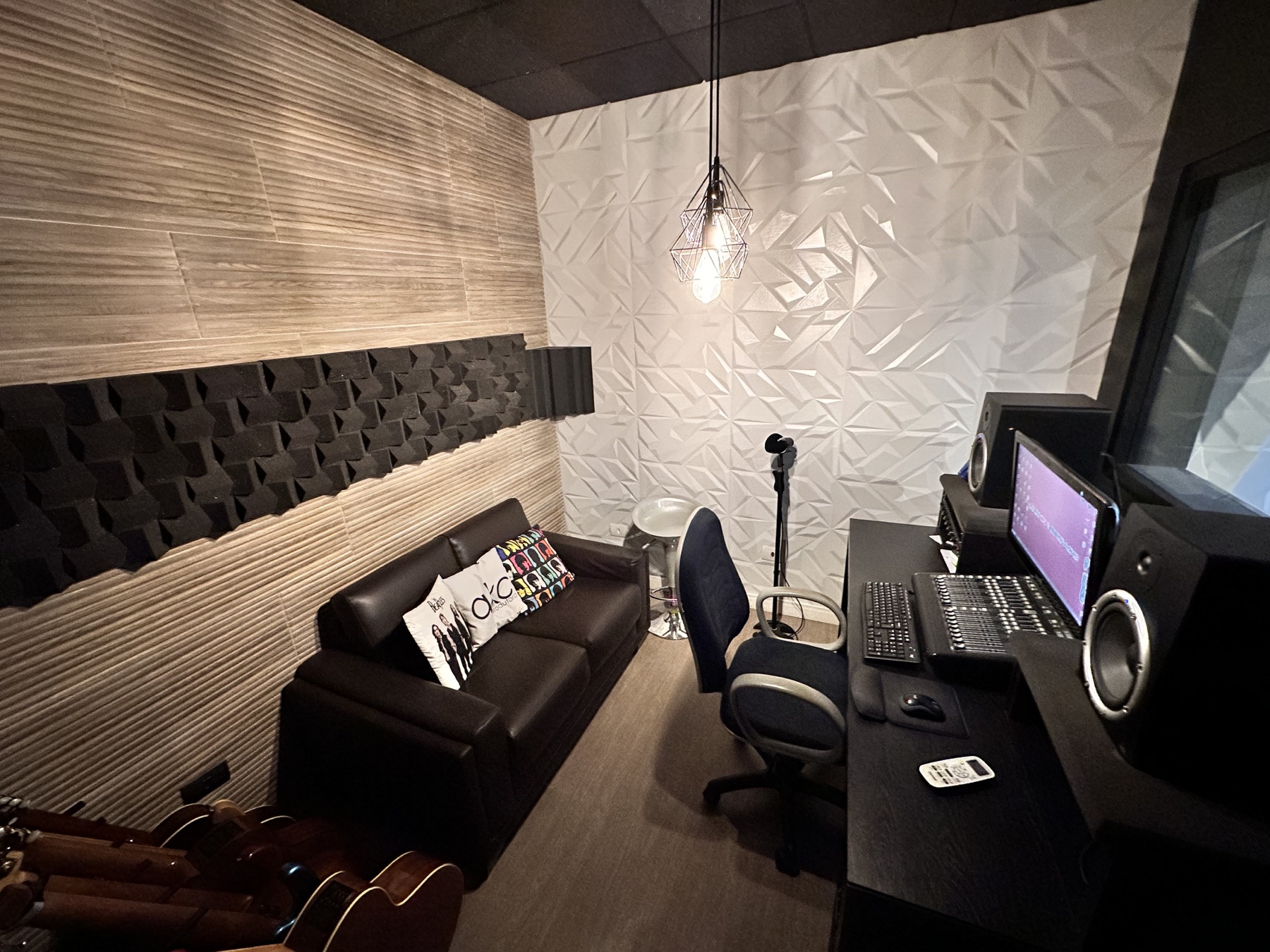 Sala de produção musical com sofá de couro, violões apoiados, mesa de gravação, monitores de áudio e parede acústica em relevo
