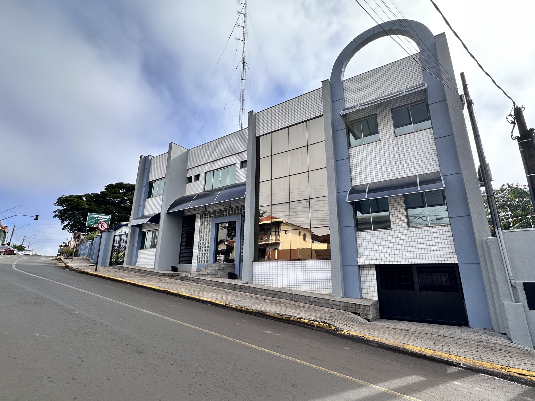 Vista lateral da sede da Rádio Graúna FM, com placa da emissora na calçada e a torre de transmissão ao fundo